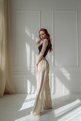 an attractive young red-haired woman in a black tank top and wide white stylish trousers. slim female model in pants, casual style