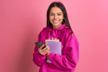 hispanic pretty woman in pink hoodie smiling holding holding notebooks and using smartphone