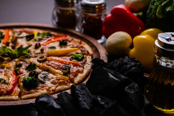 High angle shot of testy pizza along with vegetables and charcoal on wooden surface.