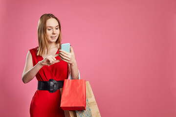 Studio shot of happy woman shopper holding shopping bags using mobile apps for online shopping on pink background. E-commerce, sale, mall discounts in applications concept.