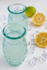 Aerial view of two glasses with lemon juice on white wooden table with half lemons, lime and ice, in vertical