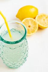 Top view of a glass with lemon juice with yellow straw and half lemons, selective focus, on white table, in vertical, with copy space
