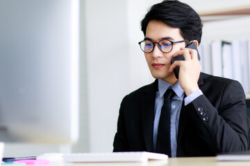 Asian handsome business man wearing eyeglasses, formal black suit with necktie, happily smiling with brace on his teeth while working and talking or communicating on mobile phone in office.