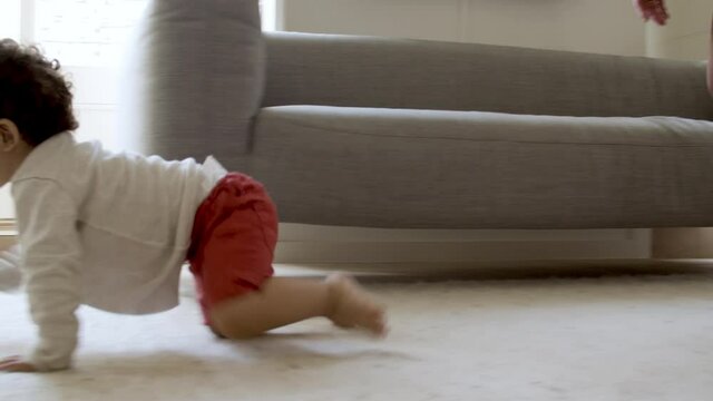 Cheerful curly-haired boy crawling on floor to ball. Cute little baby spending time with mother and father on weekend. Family, togetherness, childhood concept.