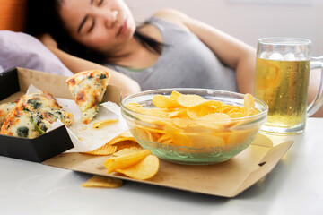 Asian woman sleep after eating junk food with pizza, potato chips and glass of beer on desk, bad...