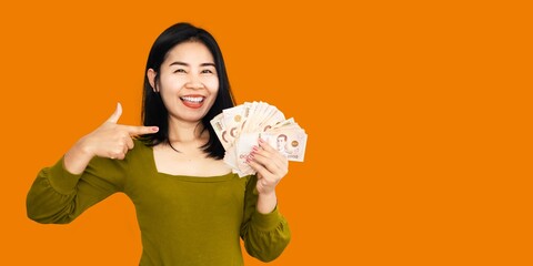 Thai woman holding money with smiling face hand pointing on banknotes over yellow background