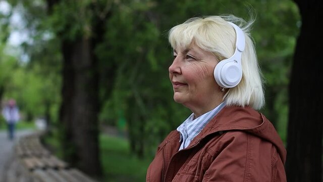 Portrait of 50 years old woman using smartphone and listening music by wireless headphones on street outdoors in city park.