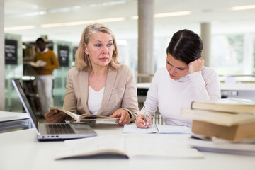 Fototapeta premium Friendly female tutor helping to diligent positive girl preparing for exam in library. High quality photo