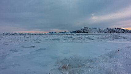 Sunset over a frozen lake. Snow and ice fragments are on the surface. Snow-capped mountain range against the backdrop of a cloudy evening sky. Baikal