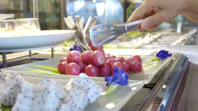 young female put fresh red grape  from salad bar to dish in hotel cafe restaurant for healthy breakfast or dinner meal with fruit in frame