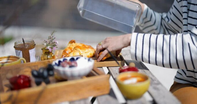 Woman having a breakfast with fresh croissants, fruits, jam and coffee outdoors, taking out croissant from a tray, close-up