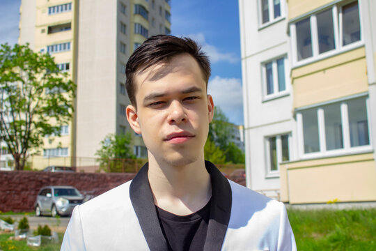 Street Portrait Of A Young Guy 17-20 Years Old In A White Suit On The Background Of A Panel House. Perhaps He Is A High School Or College Graduate, A Student Before An Exam.