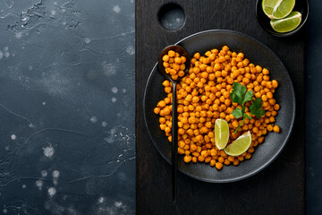 Fried chickpeas with turmeric with parsley and lime in black plate on an old black table background. Roasted spicy chickpeas or Indian chana or chole, popular snack recipe. Top view.