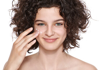 Teenager skincare. Beautiful teenage girl with gorgeous curly hair applying moisturiser face cream,...