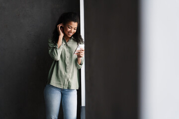 Black young woman using mobile phone while leaning on wall indoors