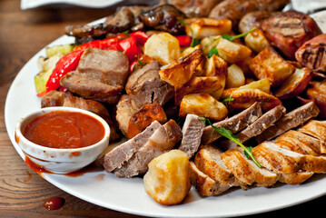 Assorted meat on a plate close up. Different types of meat and potatoes in a heap. Appetizer on a large plate with sauces.