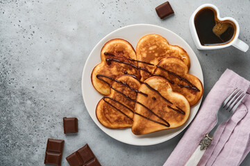 Pancakes in shape of breakfast hearts with chocolate sauce in gray ceramic plate, cup of coffee on gray concrete background. Table setting for Valentines Day breakfast. Top view copy space.