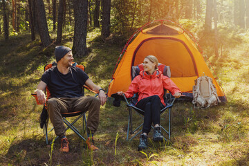 happy couple relaxing in camping chairs at campsite after hike in the forest