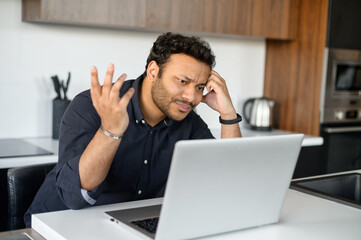 Confused indian man in smart casual shirt looking at the laptop screen, misunderstanding what happened, issues with a project or system error on the computer