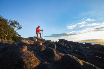Obraz premium One man in a red jacket with a trekking stick is walking on a rocky hilltop. Sunset time There is a beautiful sky.