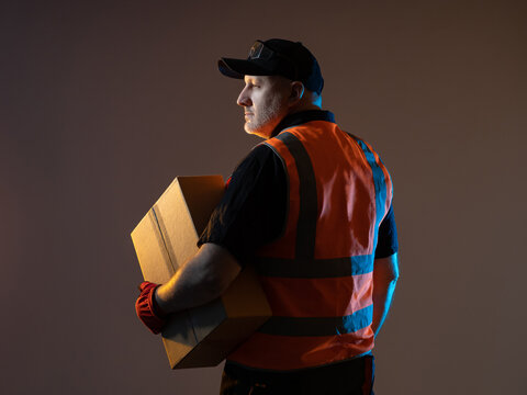 Warehouse Worker With Cardboard Box. Warehouse Worker Male. Portrait Of A Warehouse Fulfillment Employee On A Dark Background. He Is Thinking About Something. Fulfillment Center Employee.