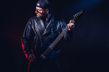 Brutal bearded Heavy metal musician in leather jacket, cap and sunglasses is playing electrical guitar. Shot in a studio on dark background