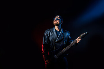 Brutal bearded Heavy metal musician in leather jacket, cap and sunglasses is playing electrical guitar. Shot in a studio on dark background