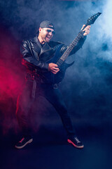 Fototapeta premium Full size photo of brutal bearded Heavy metal musician in leather jacket and sunglasses is playing electrical guitar very emotionally. Shot in a studio on dark background with smoke