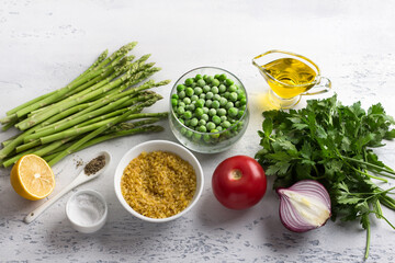 Ingredients for a middle eastern tabbouleh salad. Bulgur, fresh young green asparagus, green peas, tomato, red onion, herbs, lemon, olive oil and spices on a light blue background. Healthy vegan food