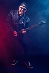 Obraz premium Full size photo of brutal bearded Heavy metal musician in leather jacket and sunglasses is playing electrical guitar very emotionally. Shot in a studio on dark background