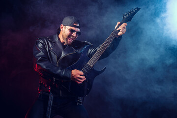 Fototapeta na wymiar Brutal bearded Heavy metal musician in leather jacket and sunglasses is playing electrical guitar. Shot in a studio on dark background with smoke