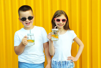 Obraz premium Cute family. Brother and sister in white t-shirts hold summer cold cocktails with orange and mint on a yellow background.