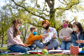 Teacher with small children sitting outdoors in city park, learning group education and coronavirus concept.