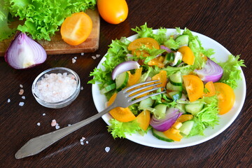 vegan salad with yellow tomatoes, red onion, cucumber and green salad leaves with pink salt and cutting board with fresh vegetables on the wooden kitchen table