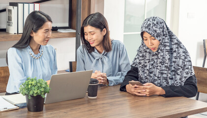 Two Asian women chatting together, and fellow Islamic women wear Hijab holding smartphones, typing messages and online, playing social networks at a company conference table together. 