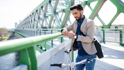 Young business man commuter with bicycle going to work outdoors in city, using smartphone.