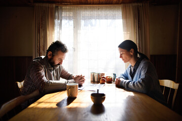 Portrait of sad poor mature couple praying at the table indoors at home, poverty concept.