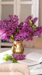 Decorative composition with a bouquet of lilacs in a gold vase on a marble table in a bright living room. Opened niga on a table with flowers. Cozy classic spring interior. Lilac bouquet. 