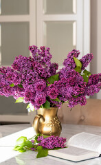Decorative composition with a bouquet of lilacs in a gold vase on a marble table in a bright living room. Opened niga on a table with flowers. Cozy classic spring interior. Lilac bouquet. 