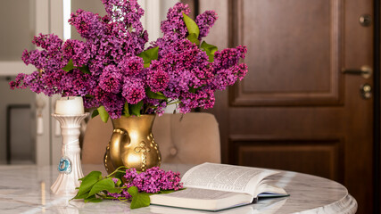 Decorative composition with a bouquet of lilacs in a gold vase on a marble table in a bright living room. Opened niga on a table with flowers. Cozy classic spring interior. Lilac bouquet. 