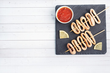 Fried squid rings on a skewer with tomato sauce and lemon. Black slate stand, white background copy...