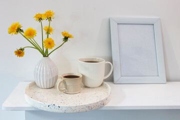 A bouquet of dandelions in a white fluted vase and two cups of different sizes with coffee on a white table.