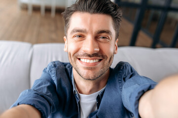 Obraz premium Young adult caucasian man takes a selfie. Portrait of attractive cheerful guy, in denim shirt, sitting on sofa, holding smartphone in hands and looking at webcam, smiling friendly
