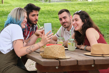 Group of happy friends looking at the smartphone having fun, drinking and eating fruit in a park. Picnic on a sunny summer day.
