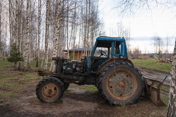 An old rusty blue tractor among the trees in the open air on a driverless farm. Agricultural machinery