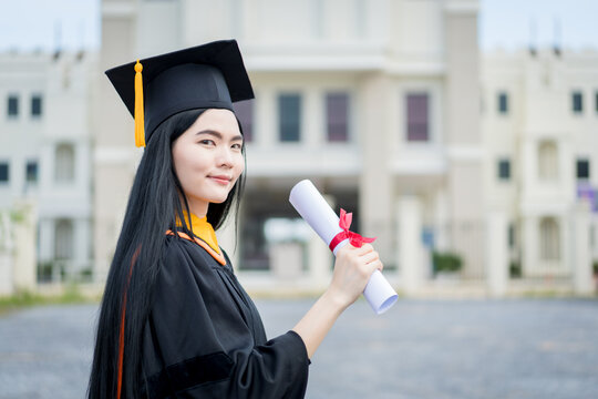 A young beautiful Asian woman university graduate in graduation gown and mortarboard holds a degree certificate stands in front of the university building after participating in college commencement