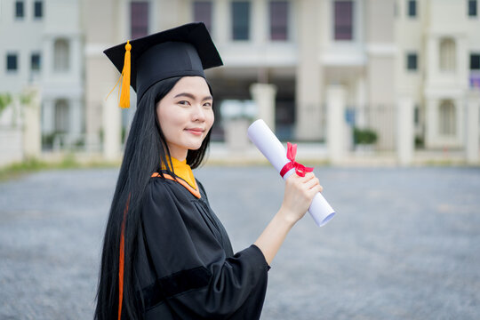A young beautiful Asian woman university graduate in graduation gown and mortarboard holds a degree certificate stands in front of the university building after participating in college commencement