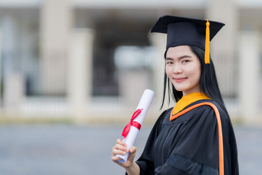 A young beautiful Asian woman university graduate in graduation gown and mortarboard holds a degree certificate stands in front of the university building after participating in college commencement