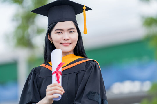 A young beautiful Asian woman university graduate in graduation gown and mortarboard holds a degree certificate stands in front of the university building after participating in college commencement