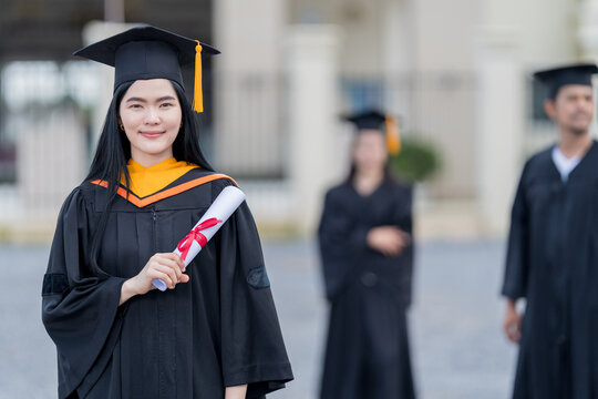 A young beautiful Asian woman university graduate in graduation gown and mortarboard holds a degree certificate stands in front of the university building after participating in college commencement